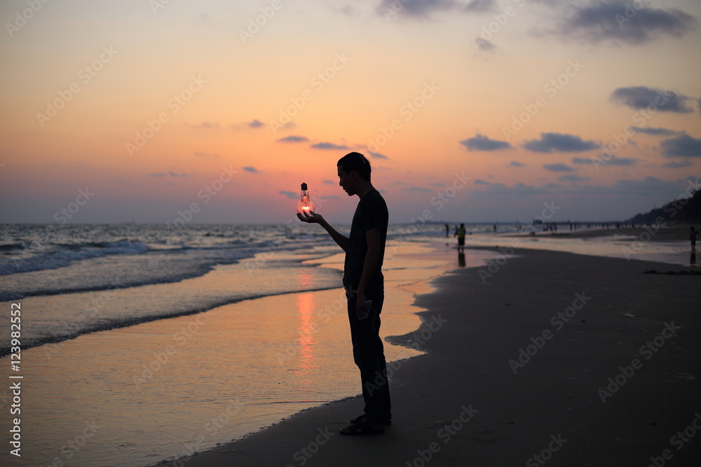 Man holds the electric light bulb and the sun on the beach at sunset