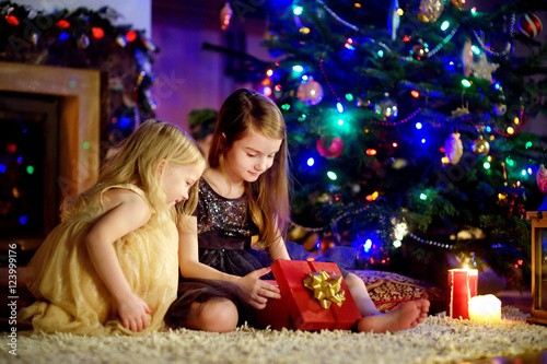 Happy little sisters opening magical Christmas gift by a fireplace