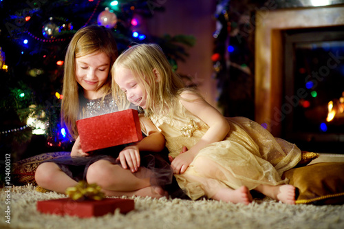 Happy little sisters opening magical Christmas gift by a fireplace