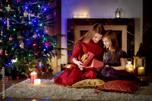 Young mother and her daughter unwrapping Christmas gifts by a fireplace in a cozy dark living room on Christmas © MNStudio