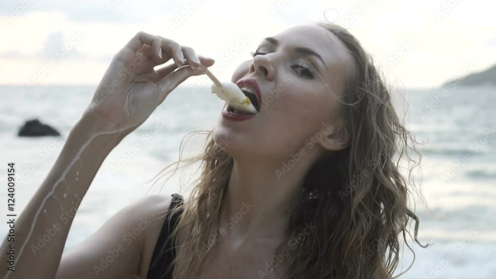 Pretty blonde happy woman in black swimsuit eating ice cream and looking into the camera at the beach over sea background - video in slow motion