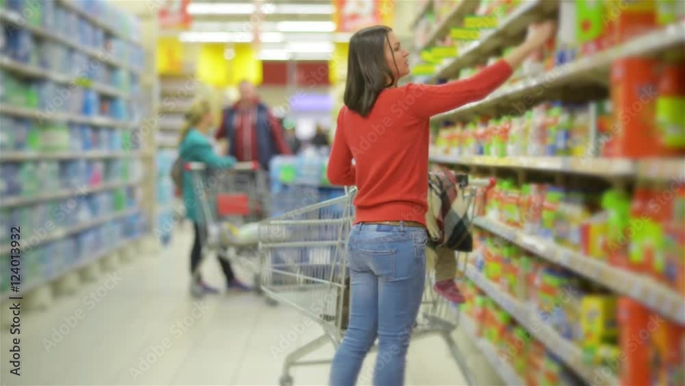 Mother and child walks along wholesale shelves and taking goods in shop trolley, beautiful young wife shopping in a supermarket
