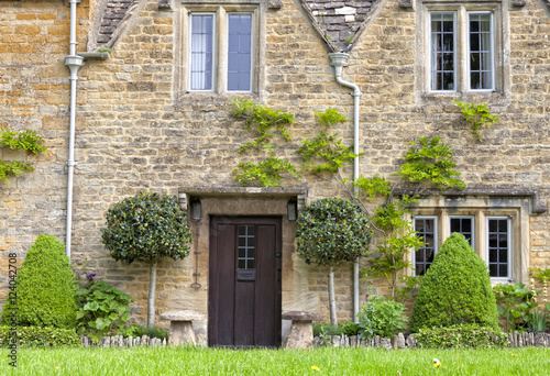 Traditional English stone cottage with topiary trees, flowers, staddle stones, grass lawn in front photo
