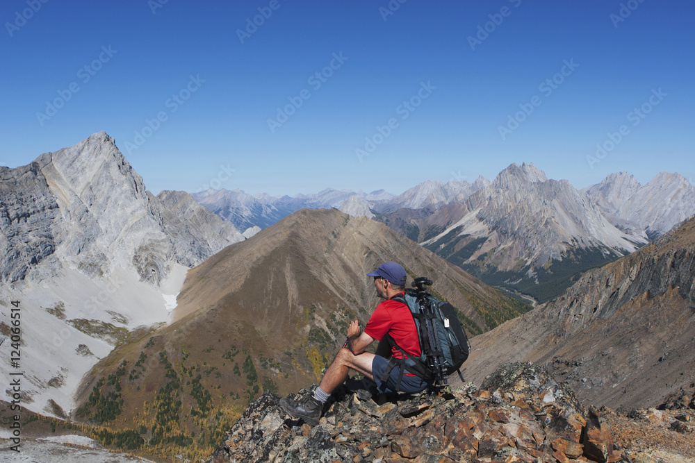 Male Hiker Sitting On Top Of Mountain Ridge Overlooking Valley And Mountains In The Distance With Blue Sky; Alberta, Canada Male Hiker Sitting On Top Of Mountain Ridge Overlooking Valley And Mountains In The Distance With Blue Sky; Alberta, Canada