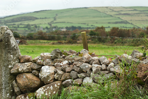 A Stone And Barbed Wire Fence With Farmland In The Background; Ireland