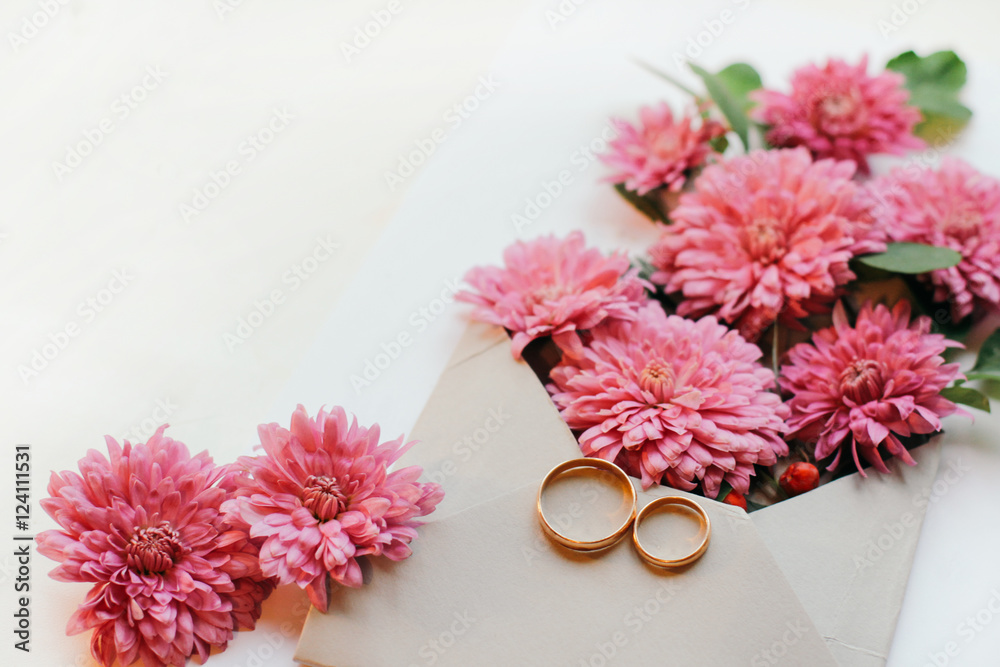 Bunch of pink chrysanthemums in an envelope and gold wedding rings. Wedding card or invitation on a white background. Season theme. Top view close-up. Bunch of pink chrysanthemums in an envelope and gold wedding rings. Wedding card or invitation on a white background. Season theme. Top view close-up.