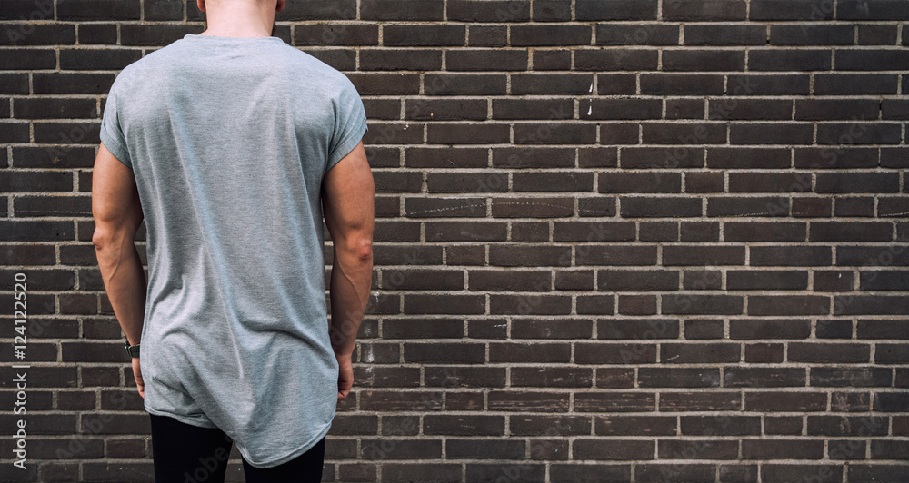 A cropped photo of a young muscular man wearing blank gray t-shirt and standing beside the dark brick wall with his back to the camera. Empty place for the logo or design. Mock up. A cropped photo of a young muscular man wearing blank gray t-shirt and standing beside the dark brick wall with his back to the camera. Empty place for the logo or design. Mock up.