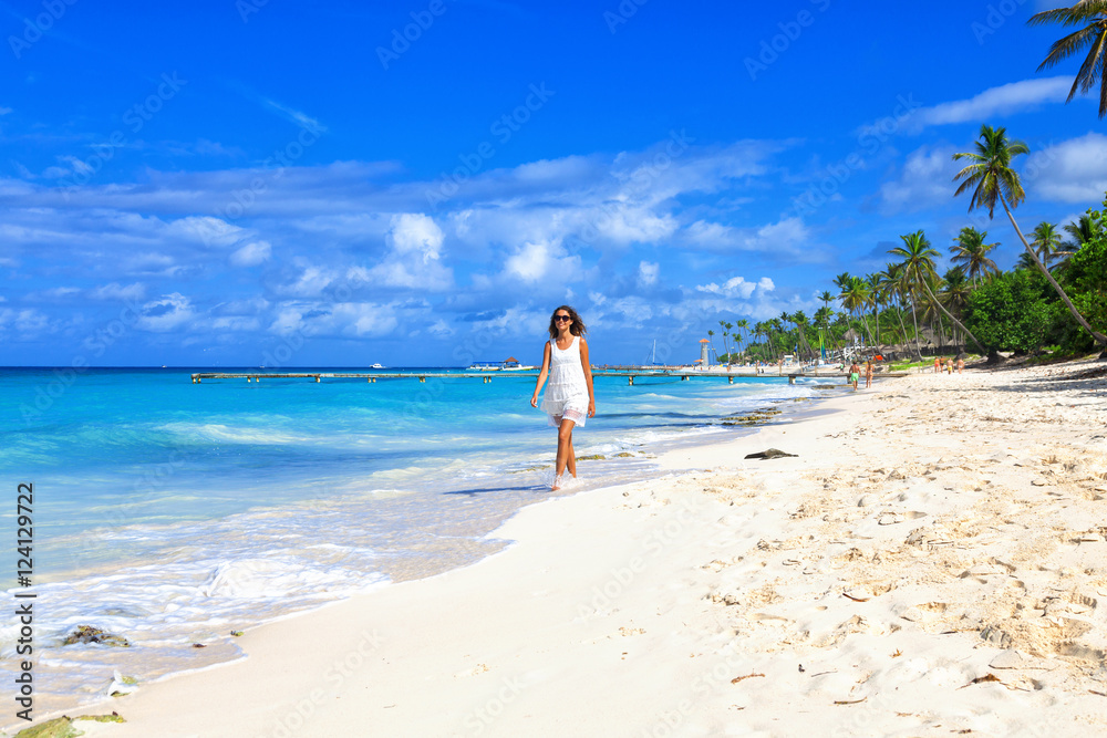 Young woman walking along white sand tropical beach. Beautiful girl in white dress on the beach. Travel and vacation. Freedom concept.
