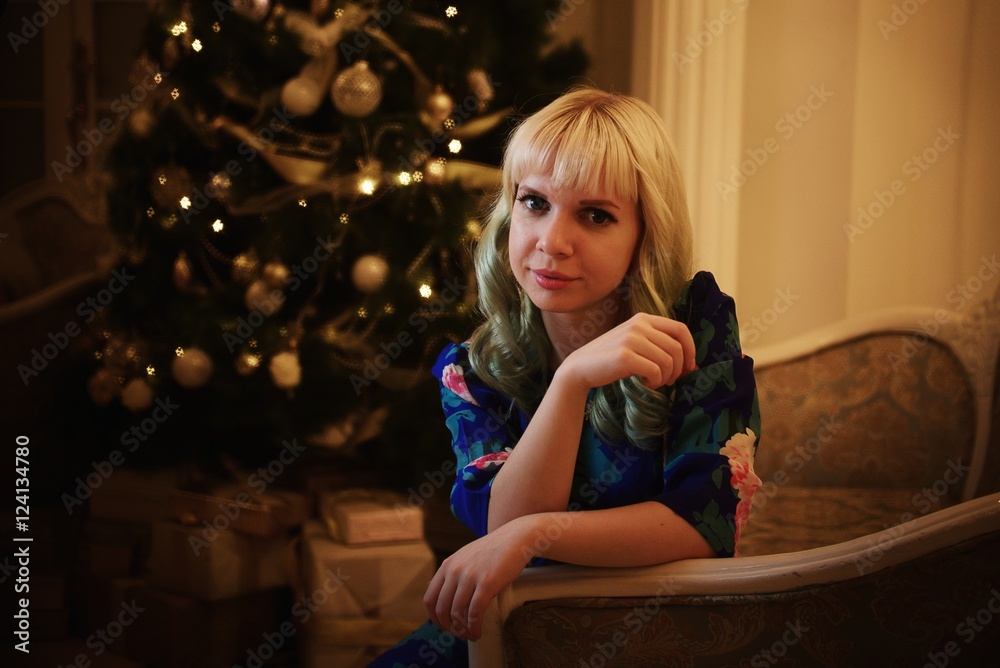 Beautiful young girl sits on sofa near Christmas tree with presents