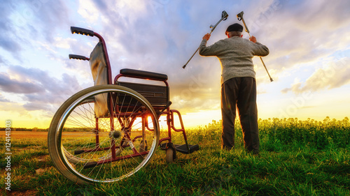 Miracle recovery: Old man gets up from wheelchair and raises hands up. Shot in a meadow. Recovery concept