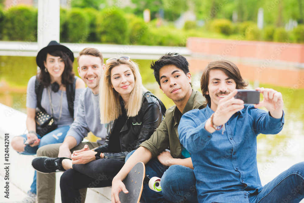 Group of young multiethnic friends sitting on a small wall, talking to each other, using a smartphone, taking a selfie , having fun - friendship, relaxing concept