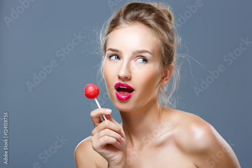 Young beautiful blonde girl holding chupa chups over grey background.
