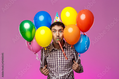 Young man celebrating birthday, holding colorful baloons over purple background.