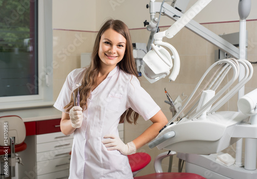 Attractive brunette female dentist with long hair, holding dental tools, smiling and looking at the camera at the modern dentist office.