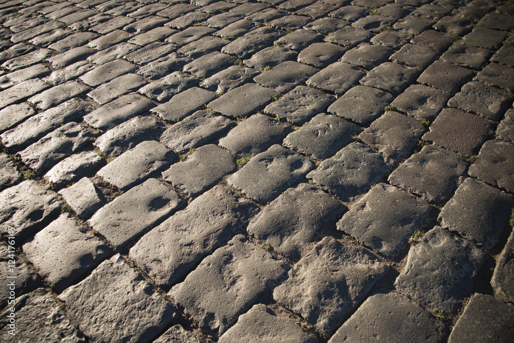 Black cobbled stone road background with reflection of light seen on the road. Black or dark grey stone pavement texture. Black cobbled stone road background with reflection of light seen on the road. Black or dark grey stone pavement texture.