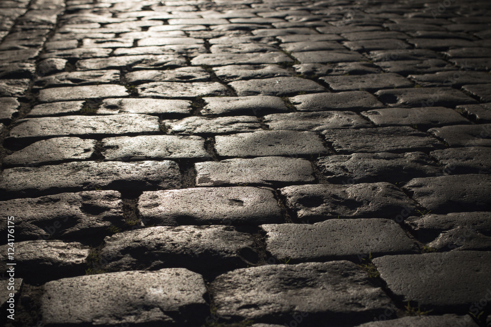 Black cobbled stone road background with reflection of light seen on the road. Black or dark grey stone pavement texture.