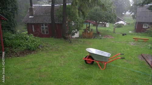 Hail and rain falling in the yard of homestead with red wooden houses and wheelbarrow photo