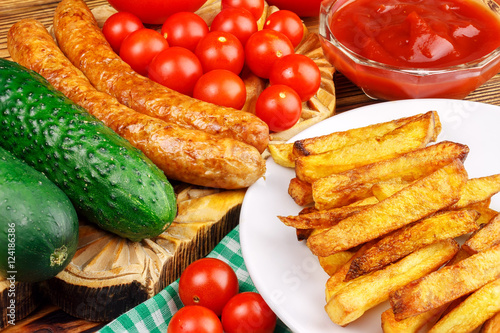 Homemade fast food, portion of french fries, ketchup, grilled sausages and cherry tomato on wooden board.