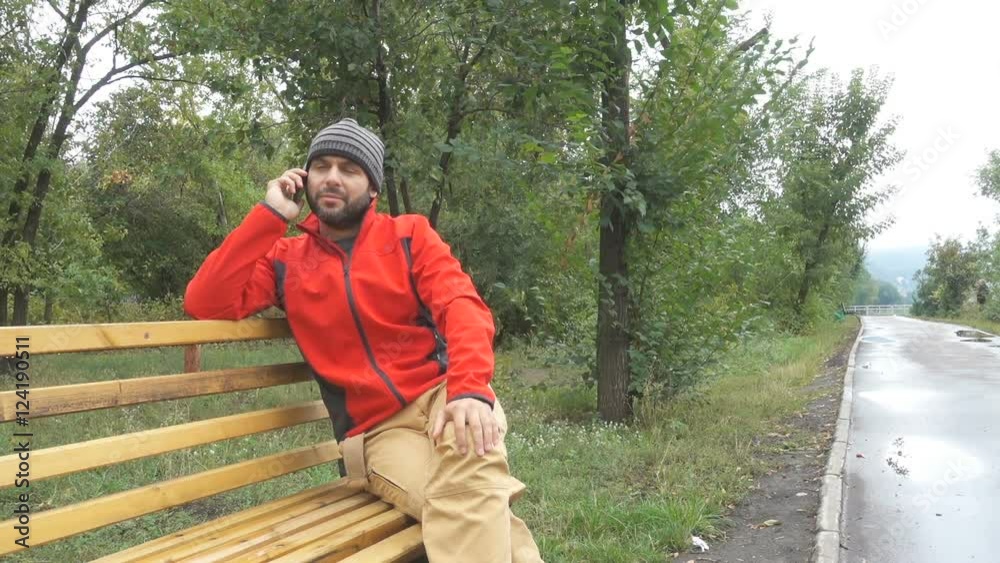 Men guy in the cap talking on phone sits on bench in autumn in city park. Men guy in the cap talking on phone sits on bench in autumn in city park.