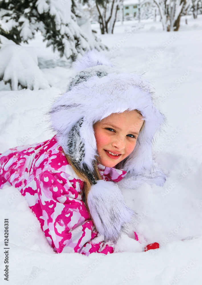 Portrait of little girl in winter clothes and warm hat with deep snow under fir tree.