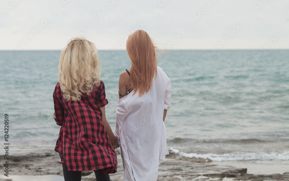 Lesbian Couple in white and red clothes near sea. Red hair and blond woman look far away on water texture Two cute girl against blue sky with clouds. Lesbian Couple in white and red clothes near sea. Red hair and blond woman look far away on water texture Two cute girl against blue sky with clouds.