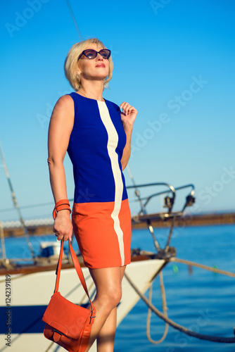 Portrait of gorgeous middle-aged blond woman in trendy dress and sunglasses holding an orange bag and looking into the distance with smile on her face. Yacht and sea on background. Outdoor shot