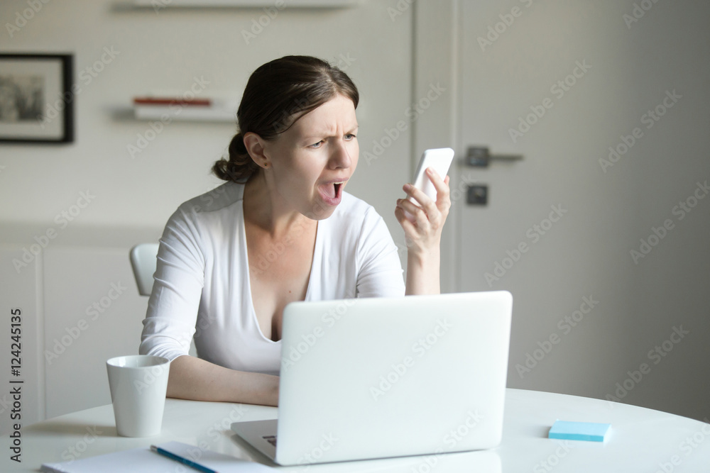 Φωτογραφία Portrait of a young woman at the desk with a laptop, looking questionnaire at the screen of the mobile she holds at her hand
