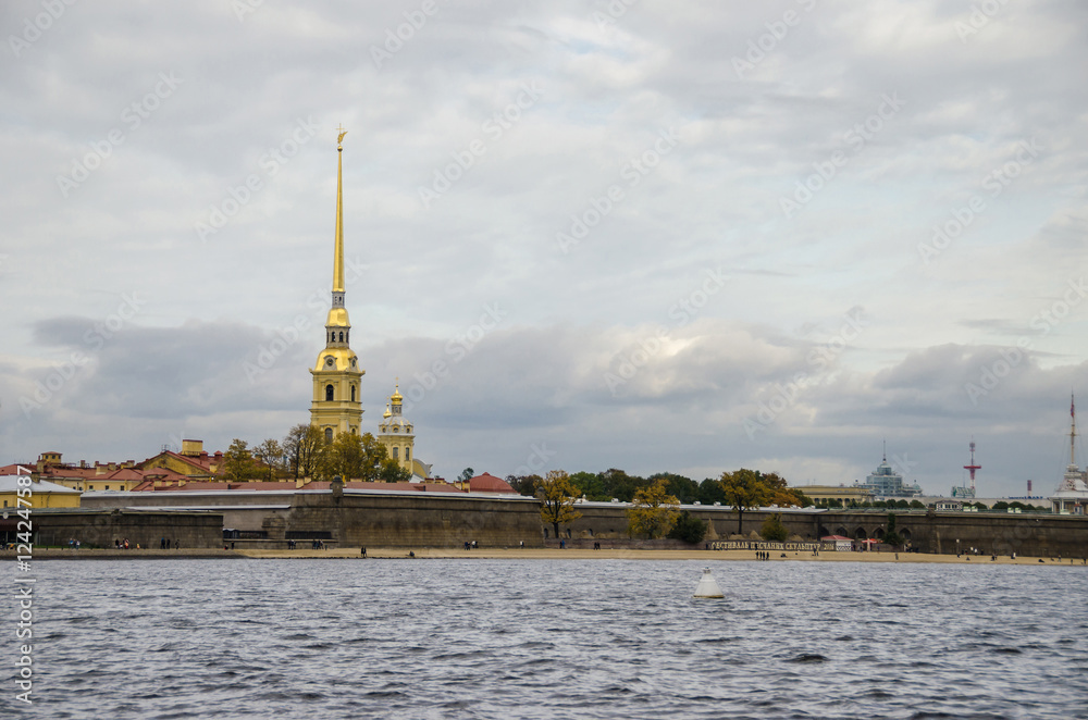 Peter and Paul Fortress and Cathedral as seen from the Spit of Vasilievsky Island with sightseeing boat and people, enjoying the gloomy weekend at the city beach, Saint Petersburg, Russia.