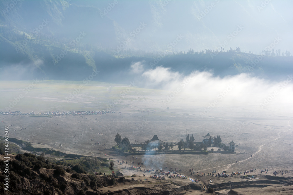 Looking down at the misty morning of caldera sea-sand plain from Mount Bromo. Tourist can be seen flocking around taking selfies, walking and horseback riding along the route to volcano crater.

