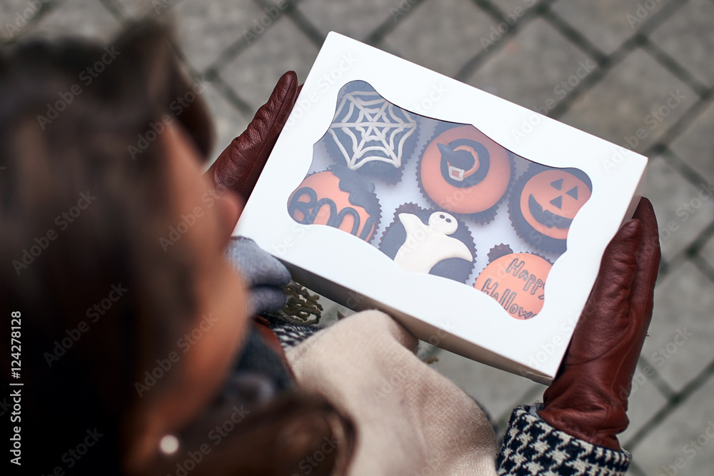 Young woman holding a colsed box full of different Halloween styled cupcakes