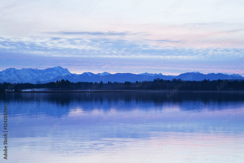 Mountain range and silhouette of a forest along the shoreline at sunset; Seeshaupt, Bavaria, Germany Mountain range and silhouette of a forest along the shoreline at sunset; Seeshaupt, Bavaria, Germany