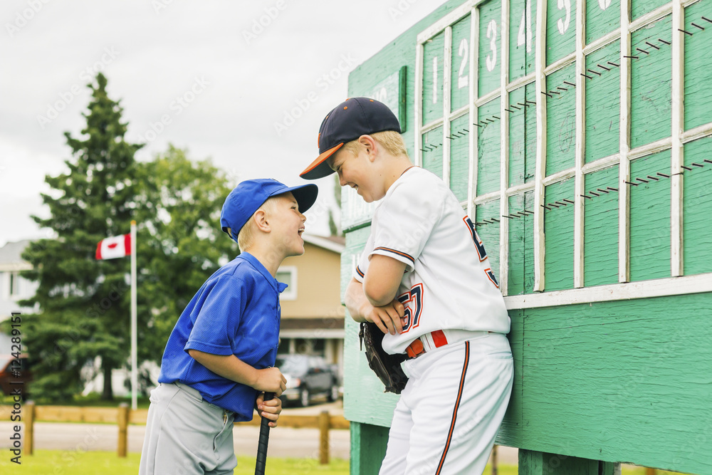 Two boys in baseball uniforms playfully argue in front of the scoreboard during a baseball game at a sports field; Fort McMurray, Alberta, Canada Two boys in baseball uniforms playfully argue in front of the scoreboard during a baseball game at a sports field; Fort McMurray, Alberta, Canada