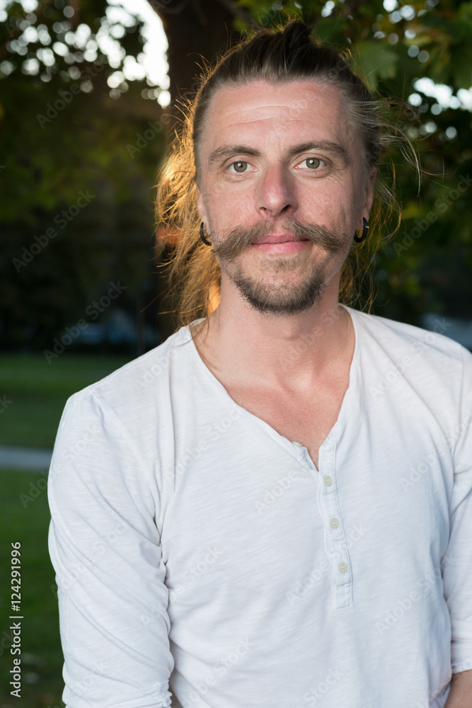 Portrait of man with long hair sitting outdoors in park while do Portrait of man with long hair sitting outdoors in park while do