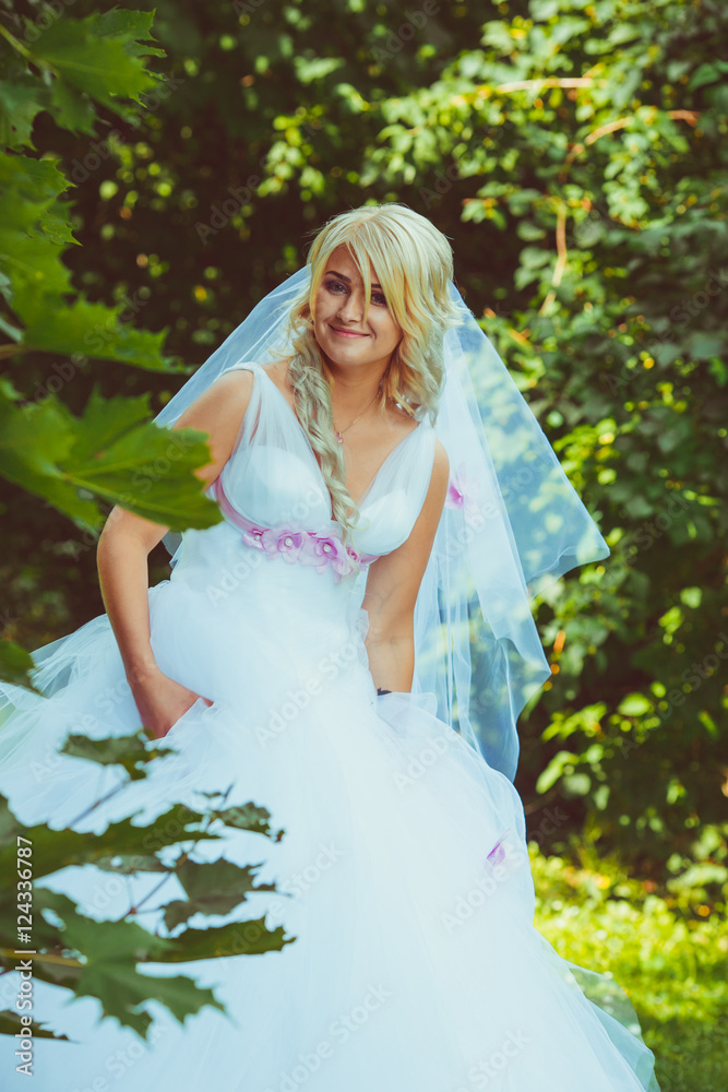 Happy blonde bride walks between the green trees holding her dre Happy blonde bride walks between the green trees holding her dre