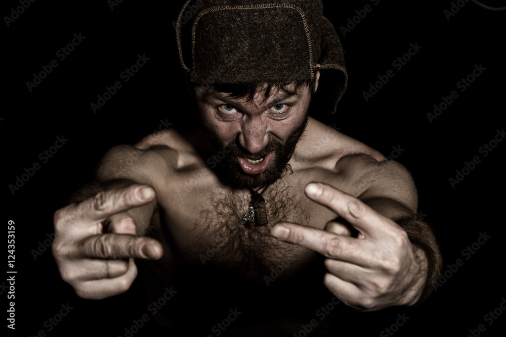 Dark portrait of scary evil sinister bearded man with smirk, shows sign of Victory. strange Russian man with a naked torso and a woolen hat