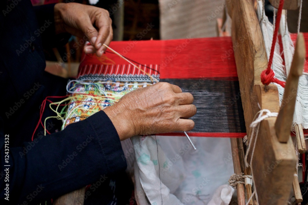 Old woman weaving red black and multi color cotton flag pattern on loom in North of Thailand