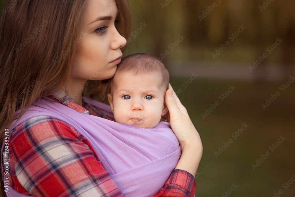 Young mother holding her little baby on the hand in sling. Walk with a child on the nature outdoor. Portrait close-up. Young mother holding her little baby on the hand in sling. Walk with a child on the nature outdoor. Portrait close-up.