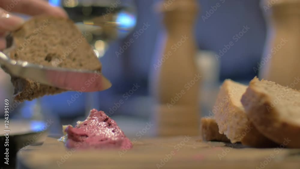 Close up view of meal table when woman hand spreading pate on wholegrain bread against background with white wine glass