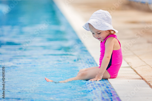 Smiling toddler girl in swimsuit and summer hat sitting at the pool, putting her legs into the water, ready to jump.  Early swimming ability. © Alina