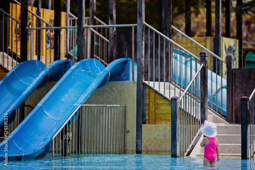 Toddler girl looking at the high slides in big aqua park with admire and fear. Summer holidays.