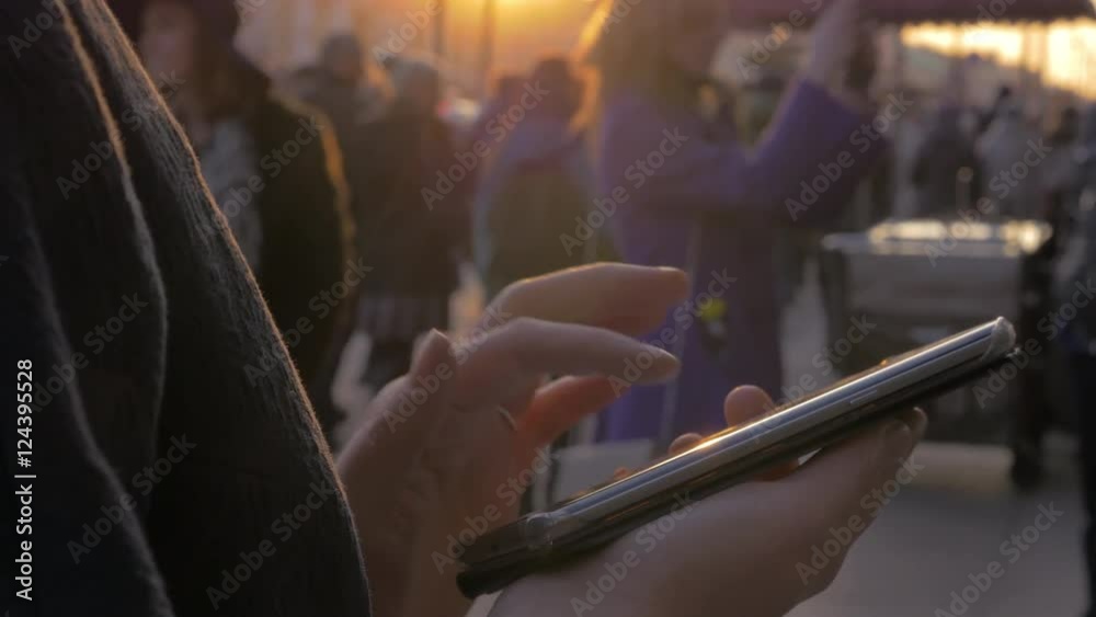 Close up view of woman hands using mobile phone against pedestrian street with a lot of people moving along against sunset light