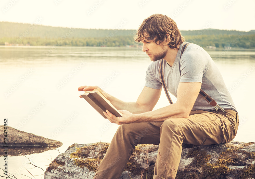 Young Man reading book outdoor with lake on background Summer va