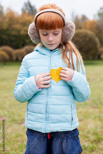 Little ginger girl in an earmuffs holding a heart shaped autumn leaf against trees