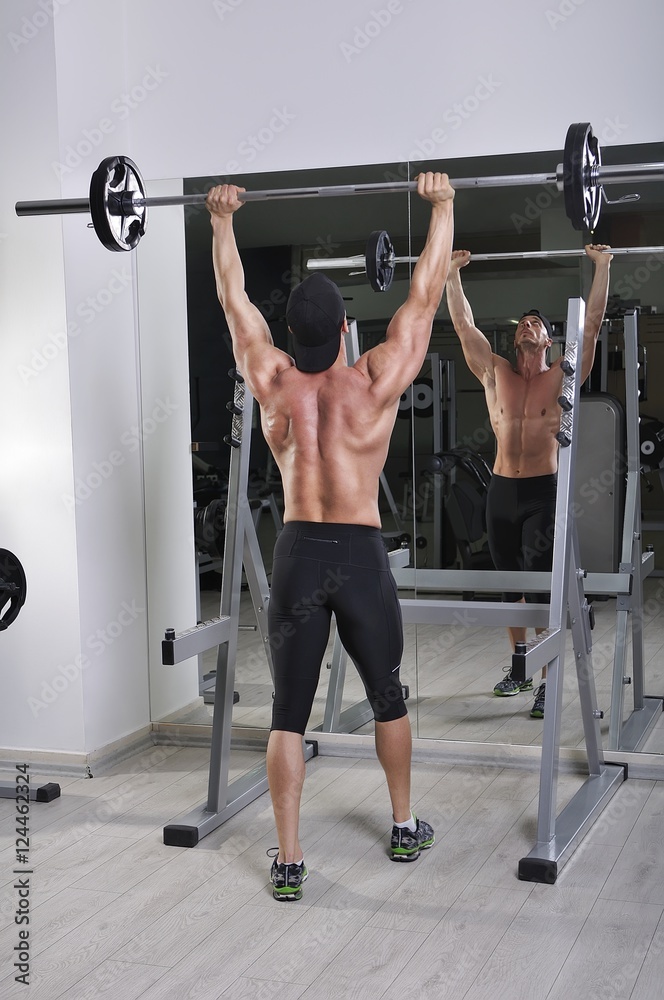 Fototapeta premium Handsome powerful athletic man doing barbell shoulder press exercise. Strong bodybuilder with perfect muscles.