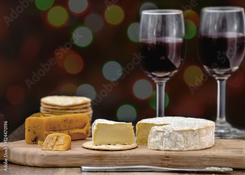 Brie and hot pepper jack cheese on wooden cheese board, accompanied by crackers and two glasses of red wine, against black background, decorated with colorful holiday lights