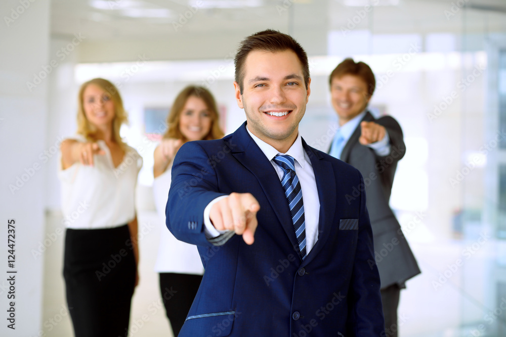 Smiling businessman in office with colleagues in the background. Pointing by finger into the camera Smiling businessman in office with colleagues in the background. Pointing by finger into the camera