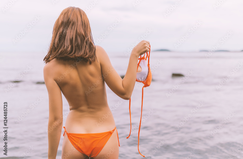 Foto Stock Rear view of a beautiful woman on a beach. She holds in her hand  the top half of her bikini. | Adobe Stock