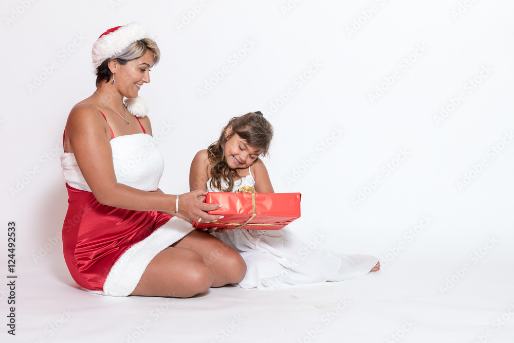 Obraz premium Tanned adult woman in Santa costume and smiling little girl are sitting on the white background. Little girl is looking at Christmas gift
