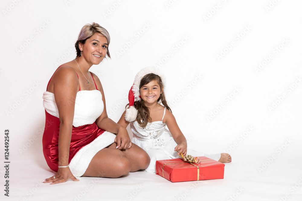 Tanned adult woman in Santa costume and smiling little girl are sitting together on the white background. They are looking at the camera Tanned adult woman in Santa costume and smiling little girl are sitting together on the white background. They are looking at the camera