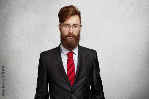 People and business. Midsection of handsome young Caucasian businessman with beard wearing rectangle glasses and formal suit, looking serious and confident, standing on gray concrete wall background